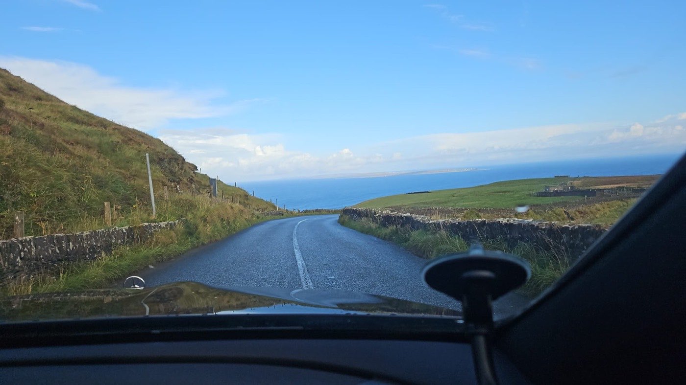 A narrow Irish coast road running down towards the Atlantic, seen through the windscreen of the car